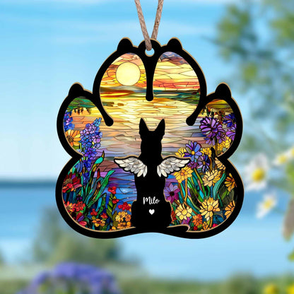 May include: A wooden paw print ornament with a stained glass design of a sunset over a lake, flowers, and a dog with wings. The dog is silhouetted and the ornament is personalized with the name "Milo".