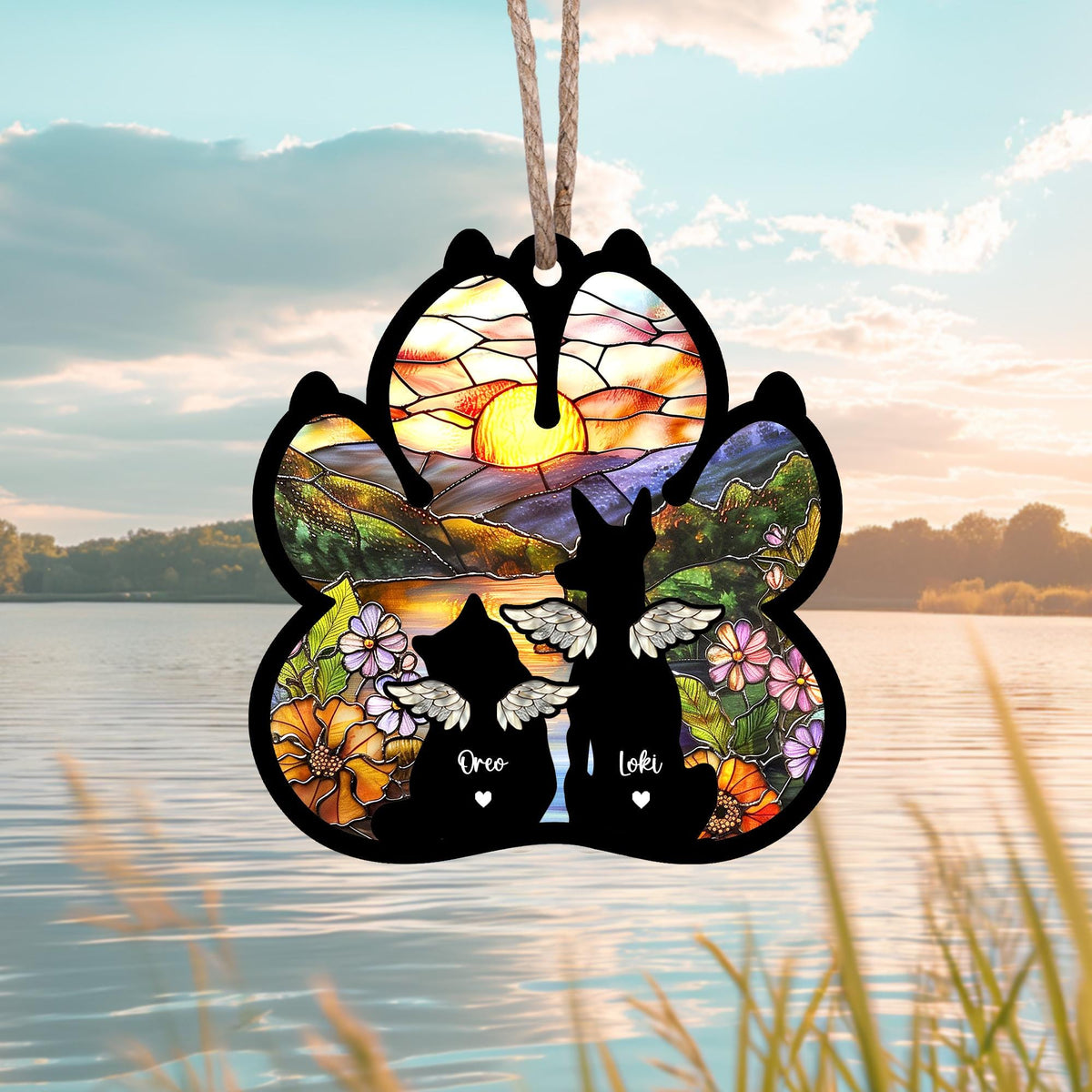 May include: A black paw print ornament with a stained glass design of a sunset over a mountain range. Two black silhouettes of cats with wings are in the center of the paw print with the names "Oreo" and "Loki" written below them.