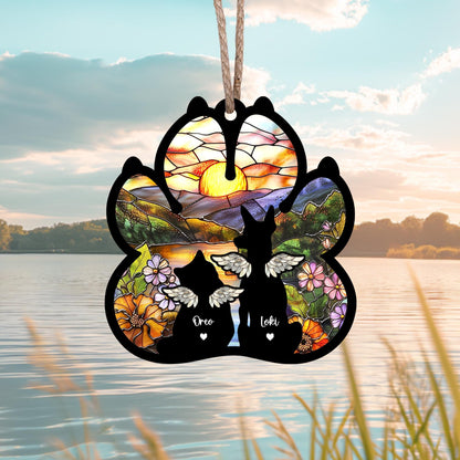 May include: A black paw print ornament with a stained glass design of a sunset over a mountain range. Two black silhouettes of cats with wings are in the center of the paw print with the names "Oreo" and "Loki" written below them.