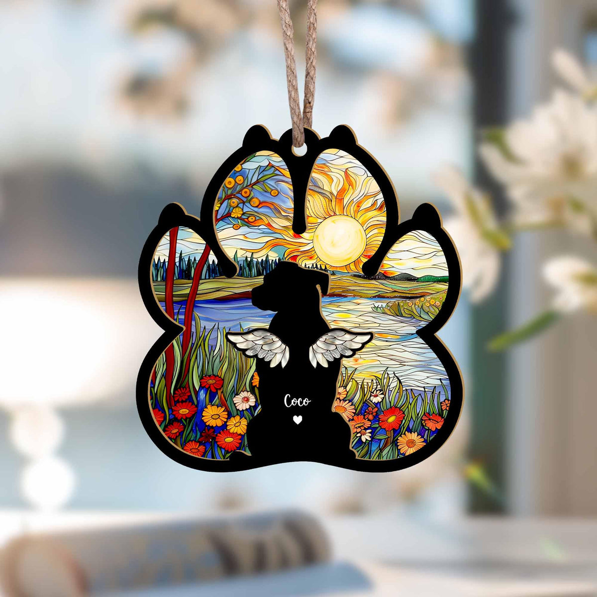 May include: A wooden paw print ornament with a stained glass design of a sunset over a lake. A black silhouette of a dog with angel wings is in the center of the ornament. The text "Coco" is written below the dog.