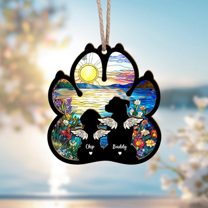 May include: A wooden paw print ornament with a stained glass design of a sunset over a lake. Two dog silhouettes are inside the paw print with angel wings and the names "Chip" and "Buddy" written below them.