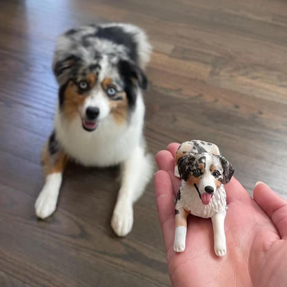 May include: A real-life Australian Shepherd dog next to a miniature figurine of the same dog. The dog has blue eyes, a white chest, and a speckled black, brown, and white coat. The figurine is held in a hand.