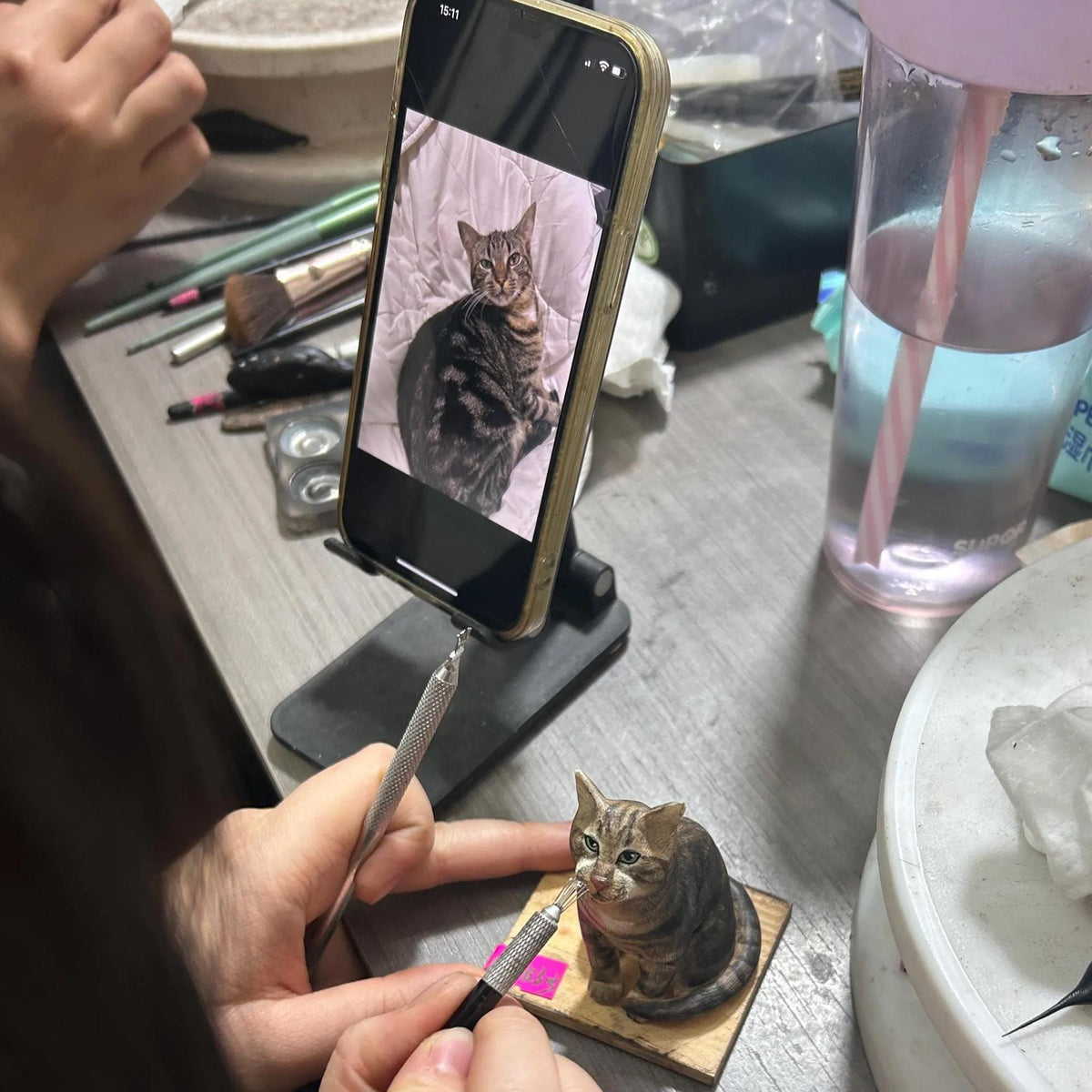May include: A detailed, miniature sculpture of a tabby cat, brown and gray, is being worked on with a stylus. The cat is on a small wooden base. A smartphone displays a photo of a similar cat. The scene is set on a work surface with art supplies.