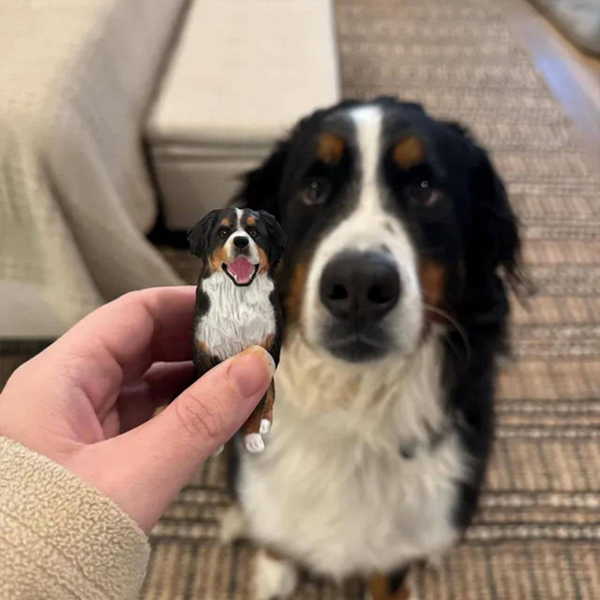 May include: A small, detailed figurine of a Bernese Mountain Dog held in front of a live Bernese Mountain Dog. The figurine has black, white, and brown markings, with a pink tongue. The larger dog has similar markings.