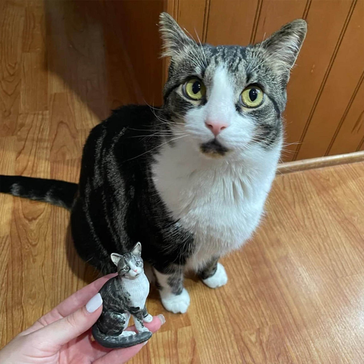 May include: A tabby cat with white markings sits next to a miniature ceramic cat figurine. The cat has green eyes and a pink nose. The figurine is a smaller replica of the cat, with similar markings.