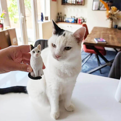 May include: A white cat with black markings sits next to a small, detailed figurine of a cat with similar markings. The figurine is held in a person's hand. Both cats have black tails and ears.