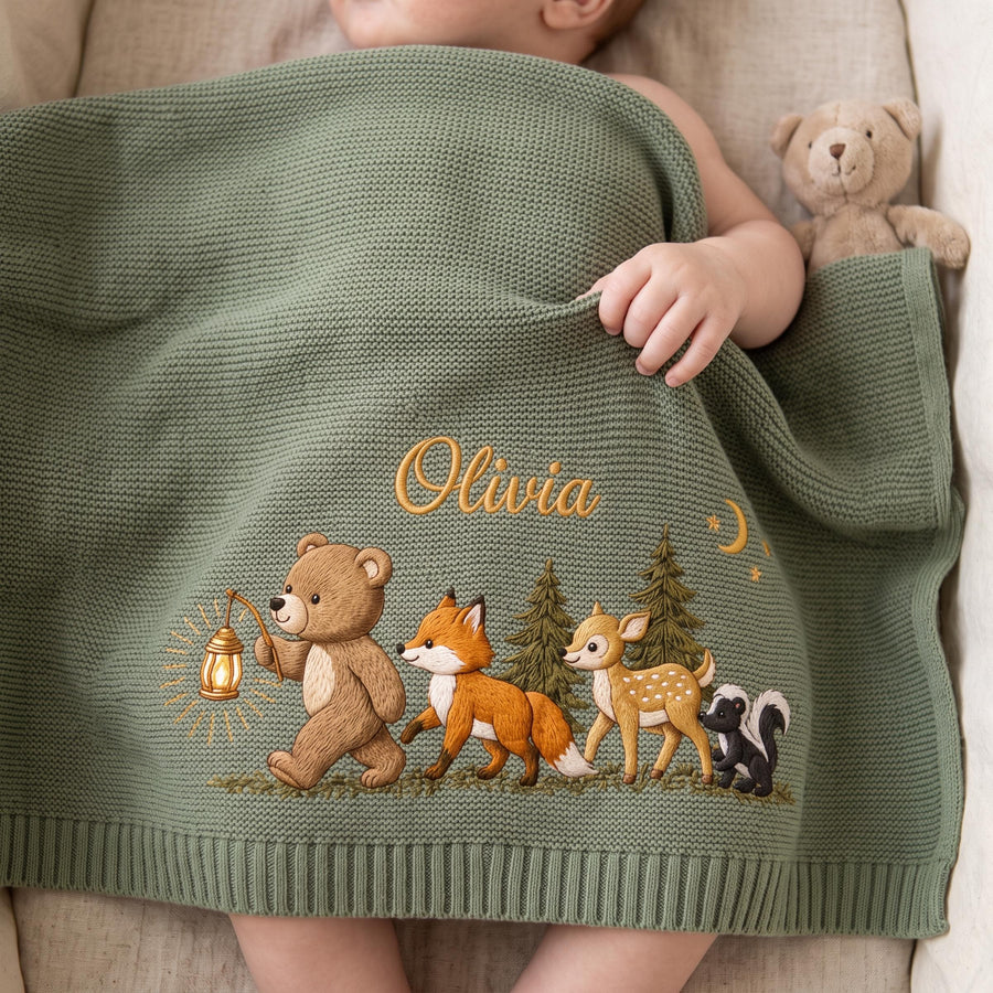 A sage green knitted baby blanket featuring the embroidered name Olivia in gold script. The design includes a brown bear holding a lantern, a fox, a deer, and a skunk walking past pine trees under a small crescent moon and stars.