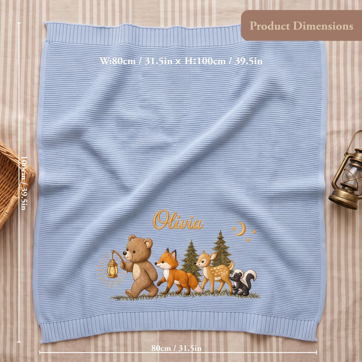 A light blue knitted blanket featuring an embroidered scene of a bear holding a lantern, a fox, a deer, and a skunk in a forest. The name Olivia is stitched in gold above the animals. The blanket measures 31.5 inches by 39.5 inches.