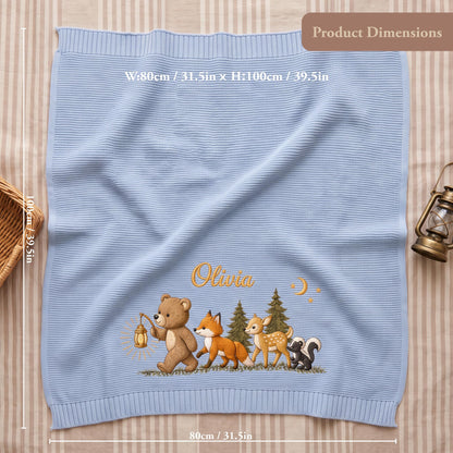 A light blue knitted blanket featuring an embroidered scene of a bear holding a lantern, a fox, a deer, and a skunk in a forest. The name Olivia is stitched in gold above the animals. The blanket measures 31.5 inches by 39.5 inches.