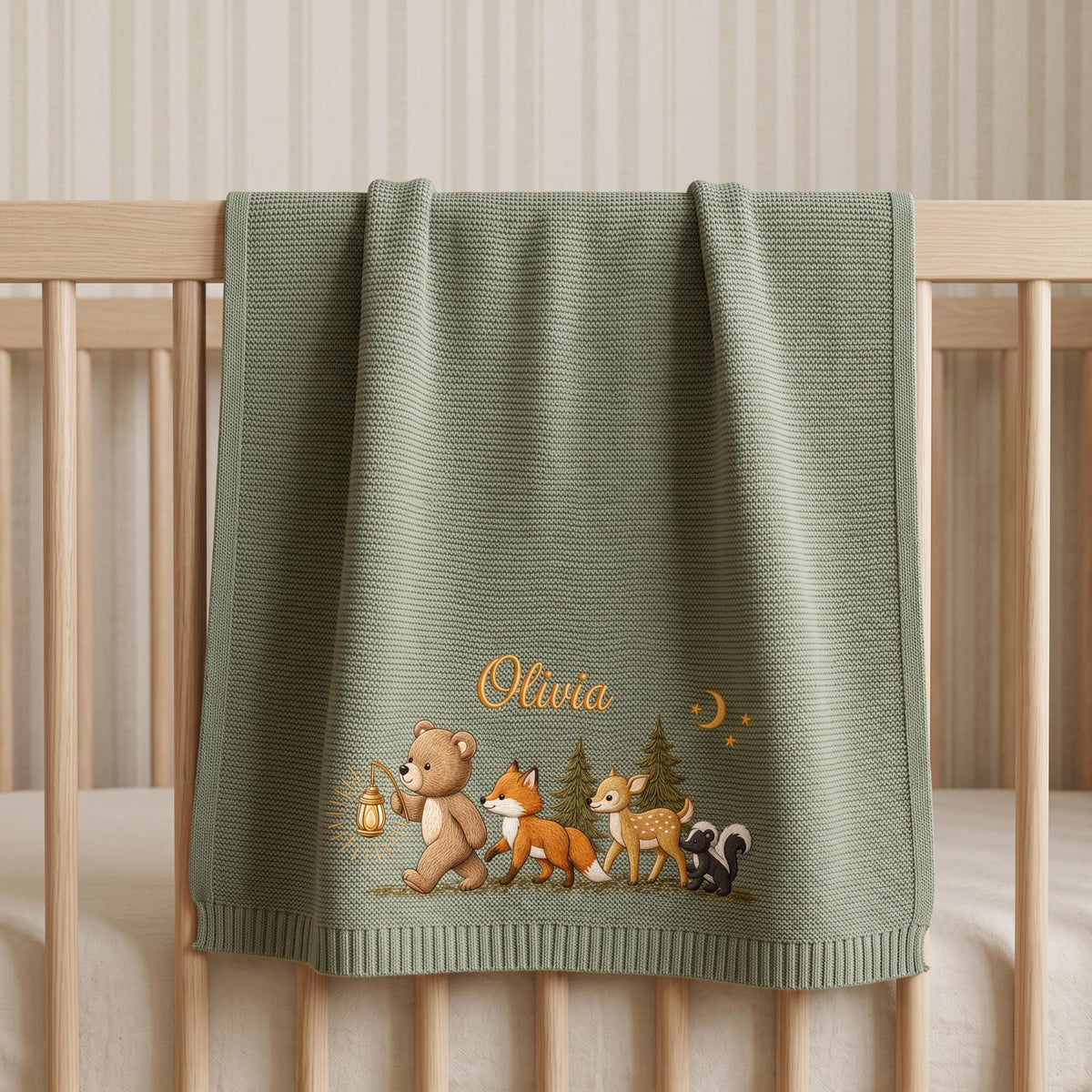 A sage green knitted baby blanket featuring an embroidered illustration of a bear holding a lantern, a fox, a deer, and a skunk near pine trees. The name Olivia is embroidered in gold script above the animals with a small crescent moon.