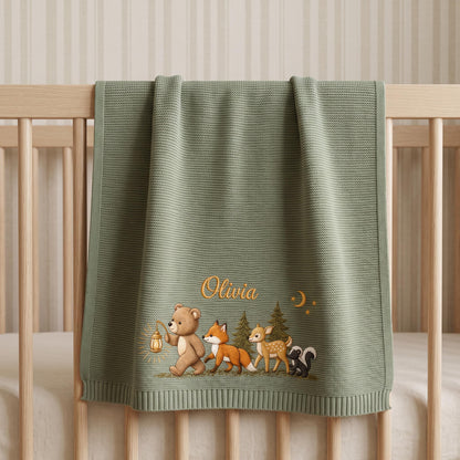 A sage green knitted baby blanket featuring an embroidered illustration of a bear holding a lantern, a fox, a deer, and a skunk near pine trees. The name Olivia is embroidered in gold script above the animals with a small crescent moon.