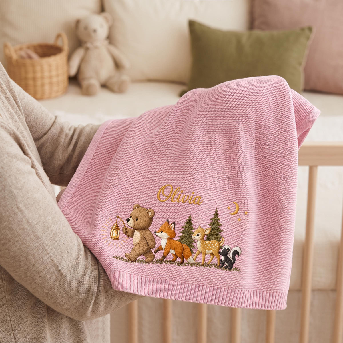A soft pink knitted baby blanket featuring an embroidered design of a bear holding a lantern, a fox, a deer, and a skunk in a forest setting. The name Olivia is stitched in gold thread above the animals, accompanied by a crescent moon.