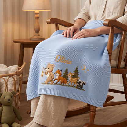 A light blue knitted baby blanket featuring an embroidered design of a bear holding a lantern, a fox, a deer, and a skunk in a forest setting. The name Olivia is embroidered in gold script above the animals, with a crescent moon.
