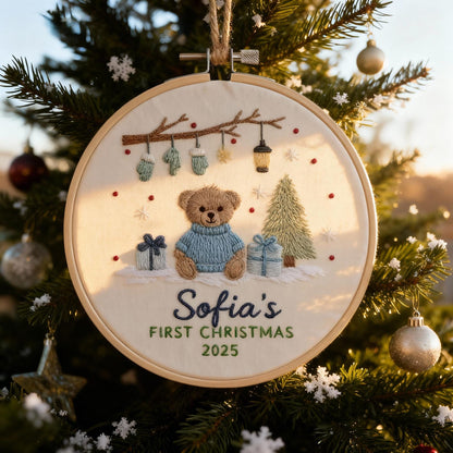 May include: Embroidered Christmas ornament featuring a teddy bear wearing a blue sweater, gifts, and a Christmas tree. The text "Sofia's FIRST CHRISTMAS 2025" is stitched below the bear. The ornament is displayed on a Christmas tree.