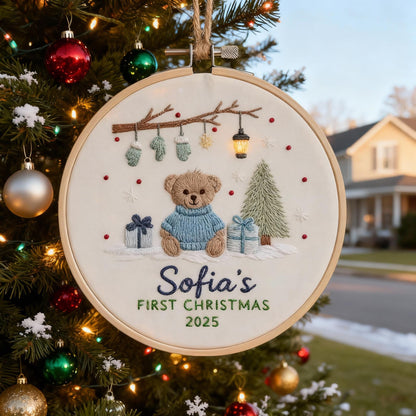 May include: Embroidered Christmas ornament in a wooden hoop. The design features a teddy bear wearing a blue sweater, gifts, a Christmas tree, and the text "Sofia's FIRST CHRISTMAS 2025". The ornament is hanging on a Christmas tree.