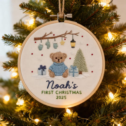 May include: Embroidered Christmas ornament featuring a teddy bear wearing a blue sweater, gifts, and a Christmas tree. The text reads "Noah's FIRST CHRISTMAS 2025". The ornament is in a wooden hoop and hangs from a twine loop.
