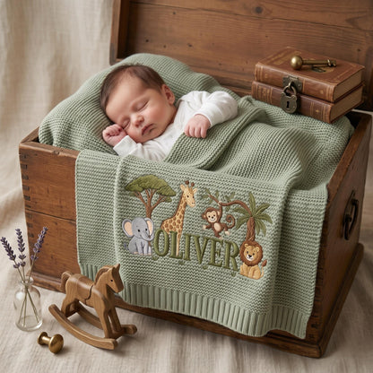 A sage green knitted baby blanket featuring embroidered safari animals including an elephant, giraffe, monkey, and lion, with the name Oliver stitched in dark green thread. The blanket is draped over a wooden chest.
