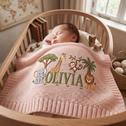 A soft, pale pink knitted baby blanket featuring an embroidered jungle scene with a gray elephant, a yellow giraffe, a brown monkey, and a lion. The name OLIVIA is stitched in large green letters across the center of the blanket.
