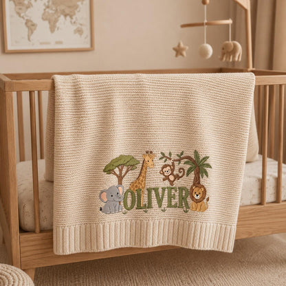 A beige knit baby blanket featuring an embroidered jungle scene with a gray elephant, a yellow giraffe, a brown monkey, and a lion. The name OLIVER is embroidered in green capital letters across the center of the blanket.