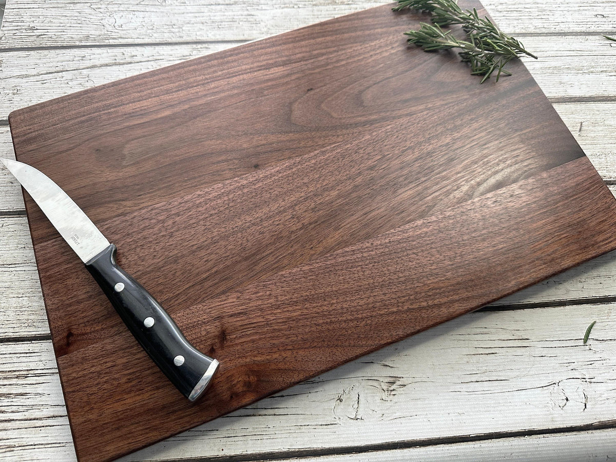 A wooden cutting board with a knife resting on it, accompanied by a sprig of rosemary, on a white wooden surface.
