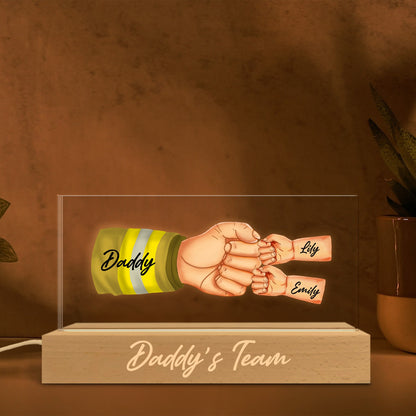 May include: A clear acrylic light-up sign with a cartoon illustration of a firefighter's hand holding the hands of two children. The firefighter's hand has a yellow and black striped sleeve with the word "Daddy" written on it. The children's hands have the names "Lily" and "Emily" written on them. The base of the sign is made of wood and has the words "Daddy's Team" written on it.