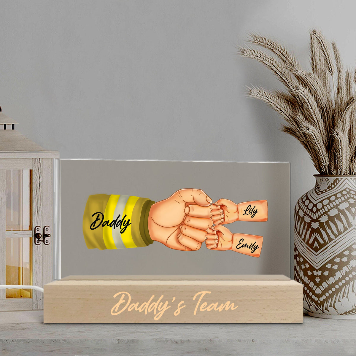 May include: A wooden light-up sign with a clear acrylic panel featuring a cartoon illustration of a firefighter's hand holding the hands of two children. The text "Daddy" is on the firefighter's sleeve and the children's names "Lily" and "Emily" are written on their arms. The wooden base of the sign reads "Daddy's Team".
