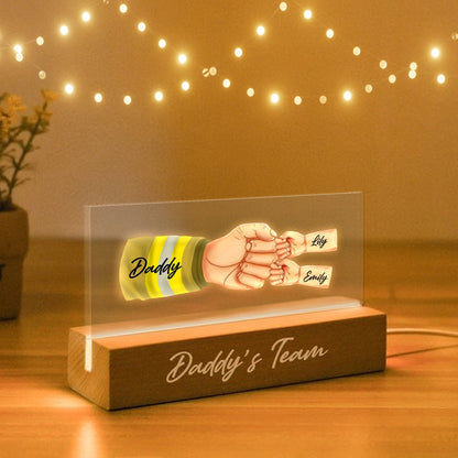 May include: A wooden base with the text "Daddy's Team" on it. A clear acrylic panel sits on top of the base and features a cartoon illustration of a hand with a firefighter's glove on it, holding the hands of two children. The children's names are written on the panel: "Lily" and "Emily".