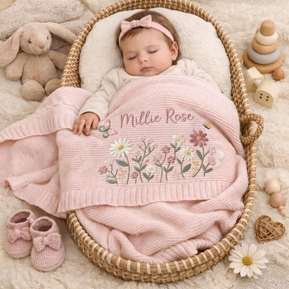 A baby wearing a light pink headband and a cream-colored long-sleeve top, sleeping under a soft pink knit blanket. The blanket features embroidered wildflowers and the name Millie Rose in cursive script. A plush bunny toy sits nearby.