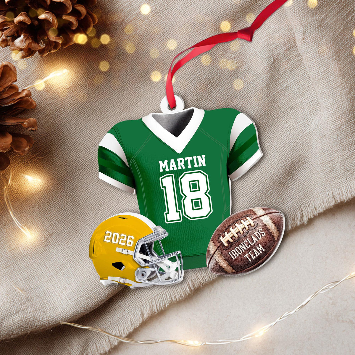 May include: A decorative ornament featuring a green football jersey with white trim, the name MARTIN, and the number 18. Below are a yellow football helmet with the text 2026 and a brown football with the text IRONCLADS TEAM, all on a red ribbon.