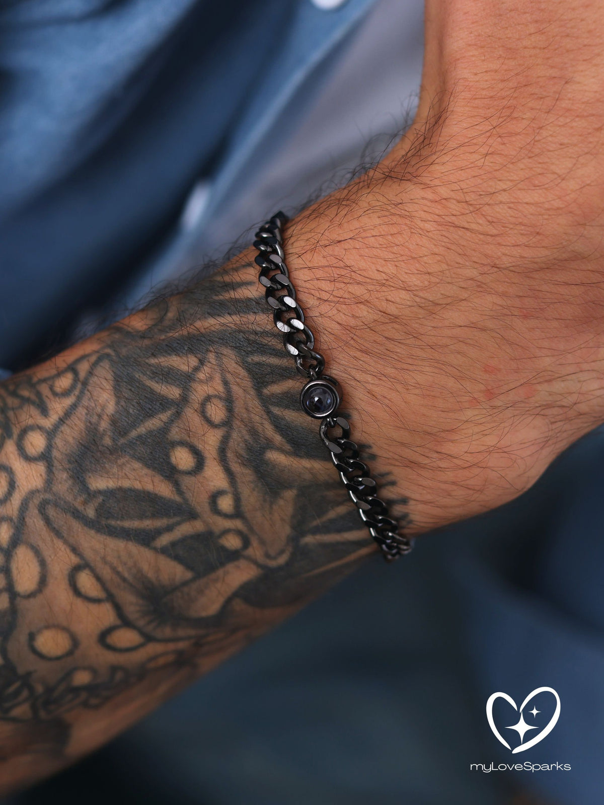 A black chain bracelet with a circular clasp. The bracelet is made of linked metal pieces and is worn on the wrist. The background is a blurred view of a blue shirt and a tattooed arm.
