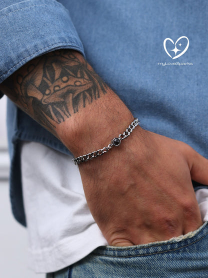 A silver curb chain bracelet with a small, dark-colored circular accent. The bracelet is worn on a wrist, with a denim shirt and white undershirt visible in the background. The brand logo "myLoveSparks" is in the upper right corner.