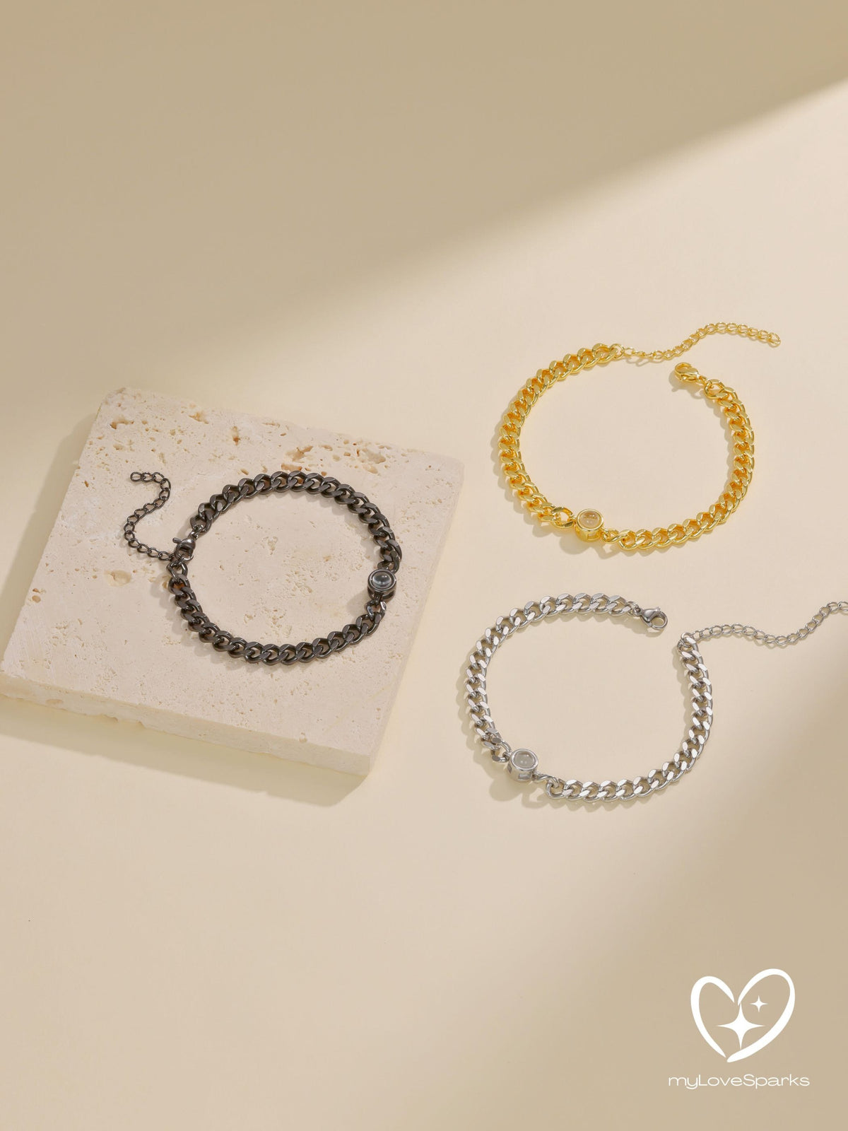Three chain link bracelets in gold, silver, and black. Each bracelet has a small, round accent piece. The bracelets are displayed on a neutral background, with one resting on a small, rectangular stone.