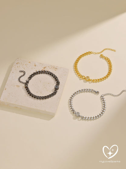 Three chain link bracelets in gold, silver, and black. Each bracelet has a small, round accent piece. The bracelets are displayed on a neutral background, with one resting on a small, rectangular stone.
