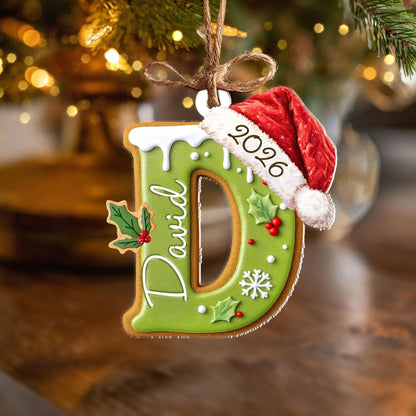 May include: A letter D shaped Christmas ornament designed to look like a green frosted cookie. It features the name David in white script, a red Santa hat with the year 2026, white snowflake accents, and green holly leaves with red berries.