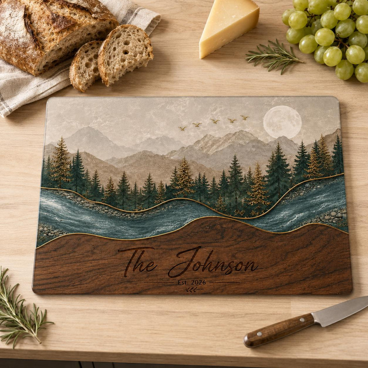 A rectangular cutting board featuring a landscape illustration of mountains, a forest, and a river under a full moon. The lower section has a wood grain pattern with the engraved text The Johnson Est. 2026. Gold lines accent the design.