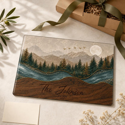 A rectangular glass cutting board featuring a landscape illustration of mountains, a forest of pine trees, a winding river, and a full moon. The lower section has a wood grain pattern with the text The Johnson Est. 2026 printed in black.