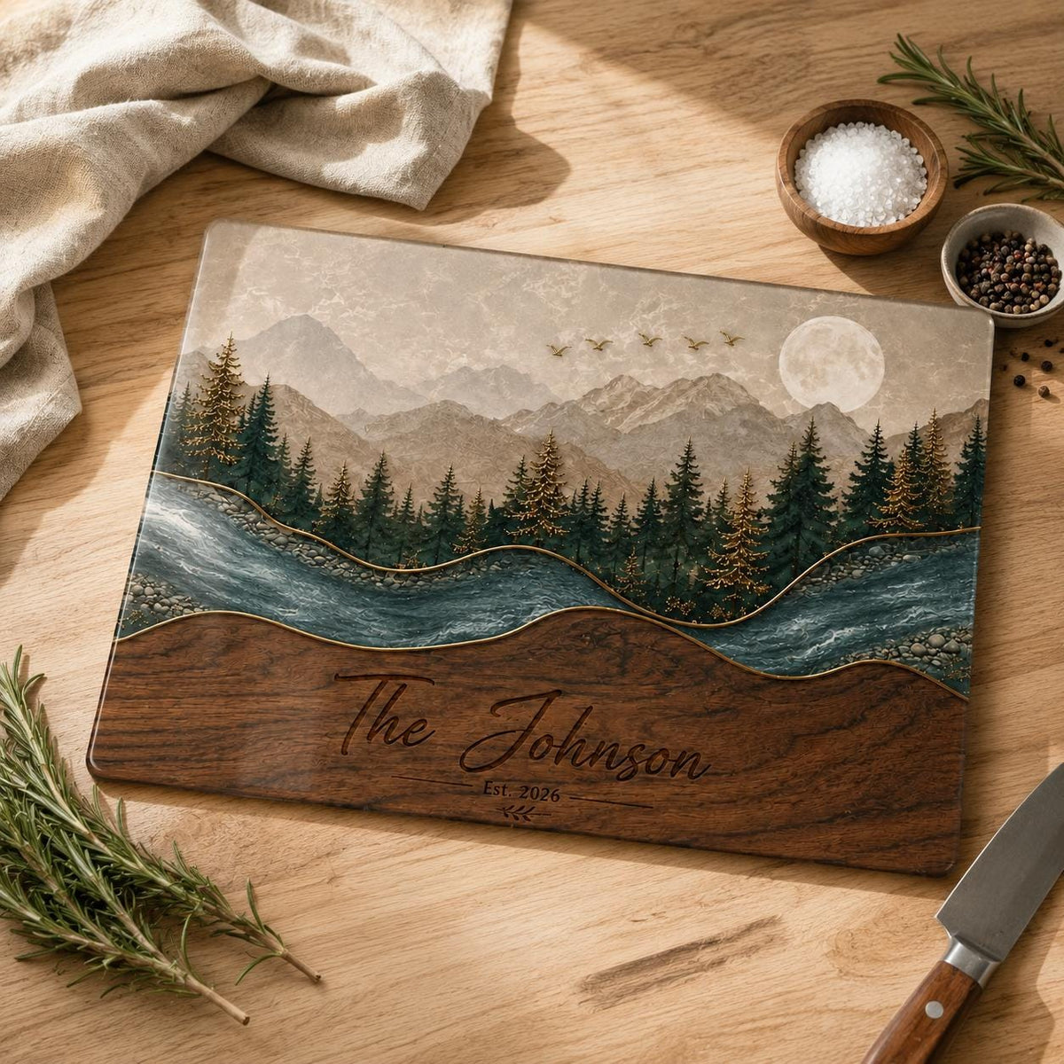 A rectangular cutting board featuring a landscape print of a forest, mountains, a river, and a full moon. The lower section is dark wood with the engraved text The Johnson Est 2026. The board has a smooth, glossy finish.
