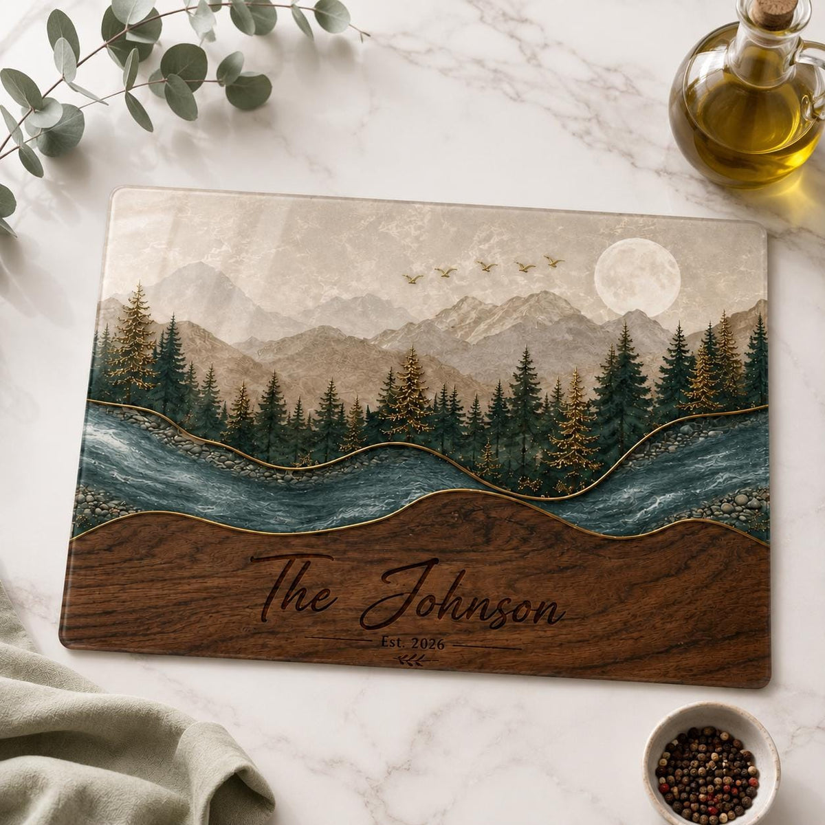 A rectangular glass cutting board featuring a printed landscape of mountains, a forest, a river, and a full moon. The bottom section has a wood grain pattern with the text The Johnson Est. 2026 printed in a dark script font.