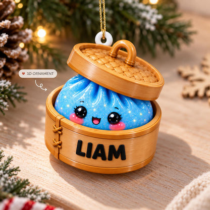A hanging ornament shaped like a bamboo steamer basket containing a blue, glittery, round dumpling character with large black eyes, pink cheeks, and a smiling mouth. The side of the steamer is personalized with the black text LIAM.