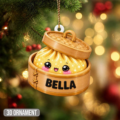 A hanging Christmas ornament shaped like a bamboo steamer basket containing a yellow dumpling with a smiling face and large black eyes. The side of the steamer is printed with the name BELLA in bold black capital letters.