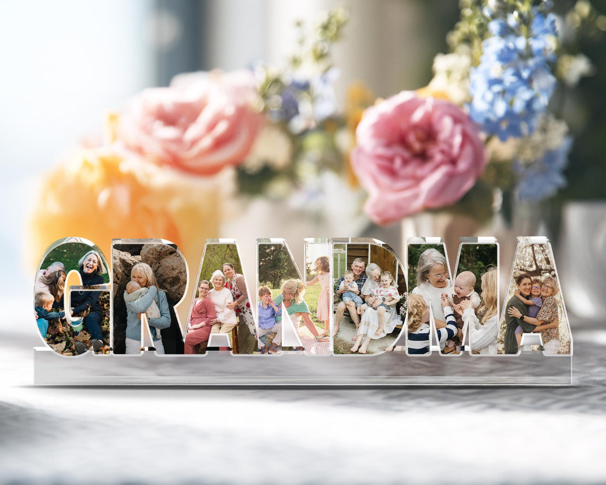 May include: A clear acrylic photo display spelling out the word "GRANDMA". Each letter contains a different family photo. The display sits on a clear, rectangular base. Soft focus background with flowers.