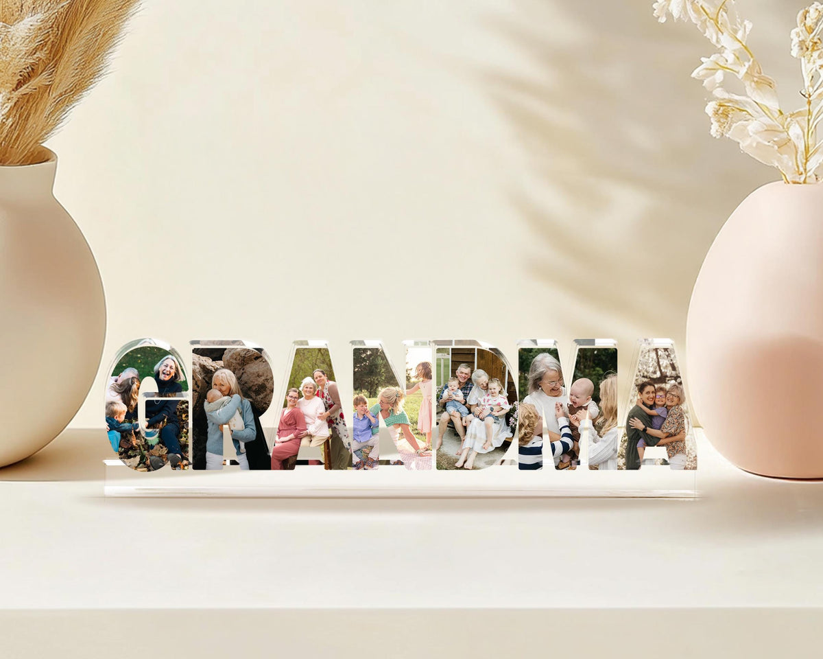 May include: A clear acrylic block spells out the word "GRANDMA" with family photos inside each letter. The block sits on a white surface, with vases and dried flowers in the background.