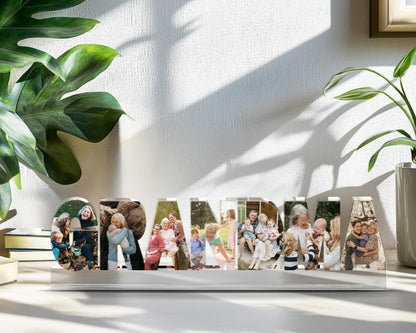 May include: A white wooden "GRANDMA" photo display. Each letter holds a different family photo. The display sits on a white surface, with green plants in the background. The photos show various family members.