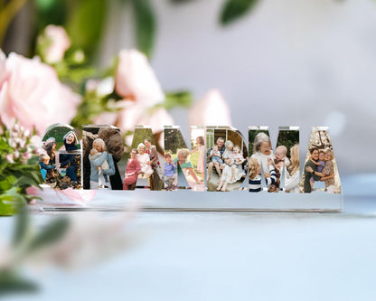 May include: A clear acrylic photo display spelling out "GRANDMA". Each letter is filled with family photos. The display sits on a clear base, with a soft-focus background of pink flowers and greenery.