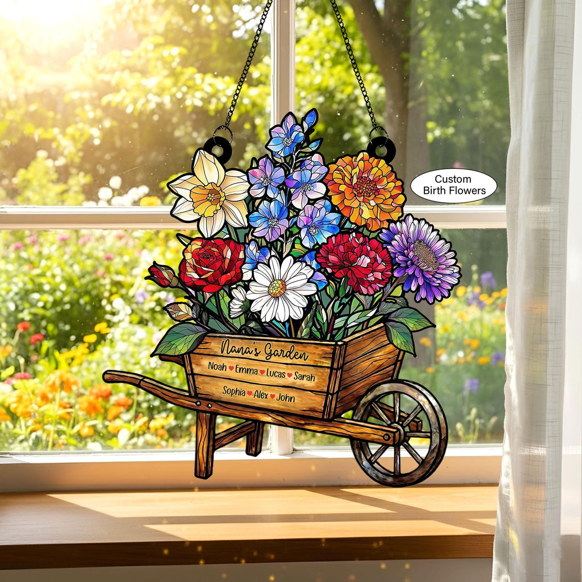 May include: A stained glass window hanging featuring a wooden wheelbarrow overflowing with colorful flowers. The wheelbarrow is labeled "Nana's Garden" with names below. The flowers include red roses, daisies, and blue and orange blooms. The words "Custom Birth Flowers" are also visible.