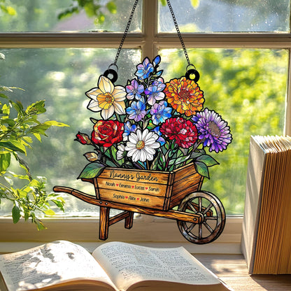 May include: A colorful floral garden sign in the shape of a wooden wheelbarrow, hanging in front of a window. The sign is filled with various flowers and the text "Nanny's Garden" with names below. The sign is suspended by a black chain.
