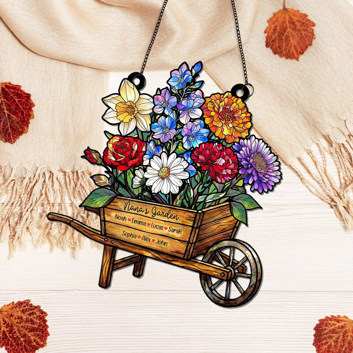 May include: A decorative wooden wheelbarrow filled with colorful flowers, including red roses, white daisies, and purple asters. The wheelbarrow has the text "Nana's Garden" and several names. The piece hangs from a black chain.