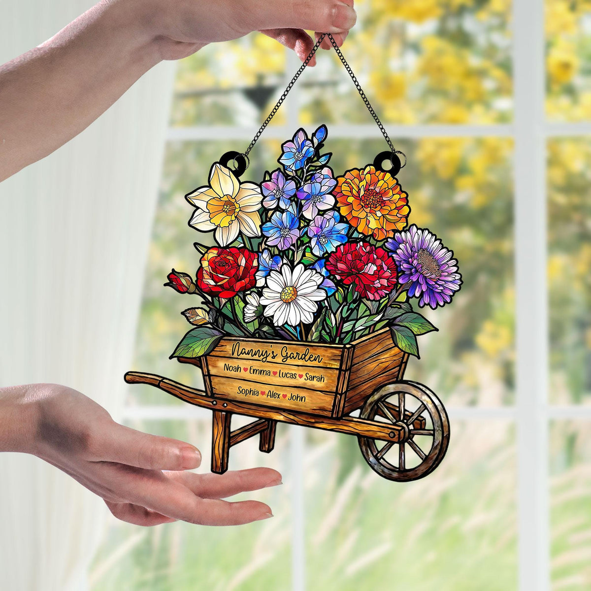 May include: A wooden wheelbarrow filled with colorful flowers, including roses, daisies, and dahlias. The text "Nanny's Garden" is above a list of names. The wheelbarrow is suspended by a chain.