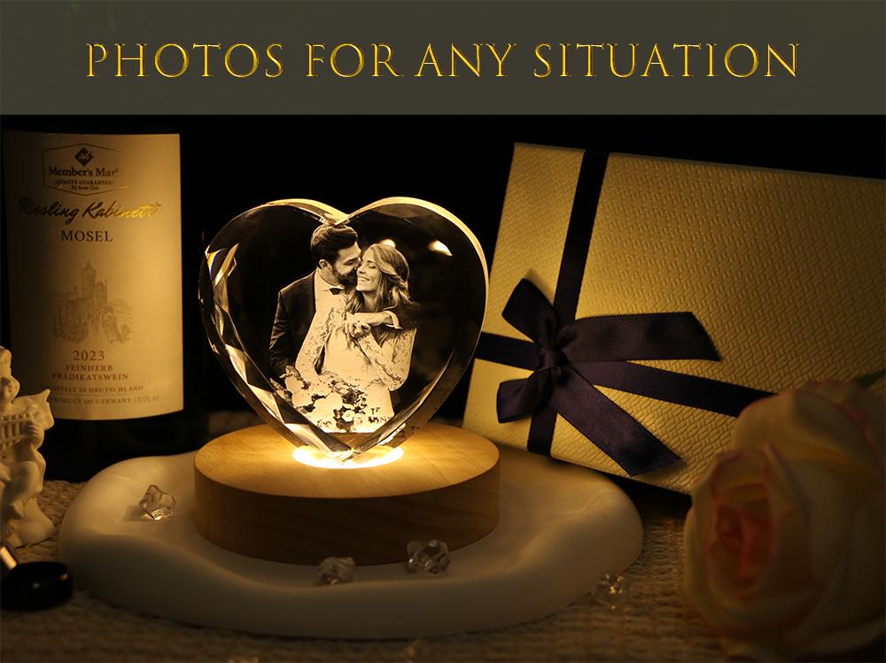 A heart-shaped glass block featuring a laser-etched photograph of a couple, illuminated by a round wooden light base. The text "PHOTOS FOR ANY SITUATION" appears at the top. A gift box with a dark blue ribbon sits to the right.