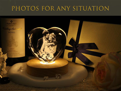 A heart-shaped glass block featuring a laser-etched photograph of a couple, illuminated by a round wooden light base. The text "PHOTOS FOR ANY SITUATION" appears at the top. A gift box with a dark blue ribbon sits to the right.